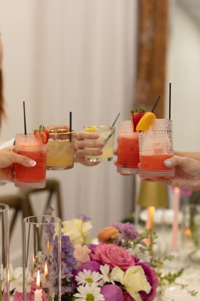 Friends toast with colorful cocktails at a floral-filled table – fun reception celebration ideas for weddings.