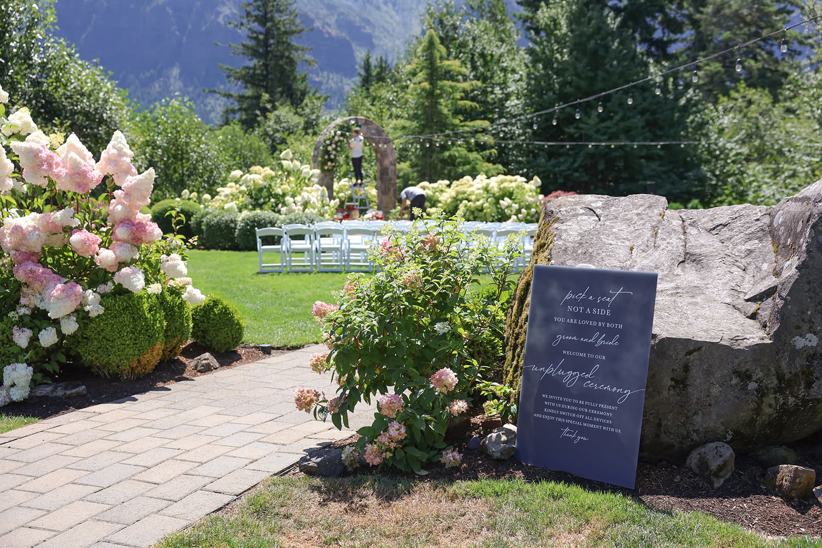 Garden ceremony setup at Cape Horn Estate with white chairs, floral arch, and unplugged wedding sign nestled in summer blooms.