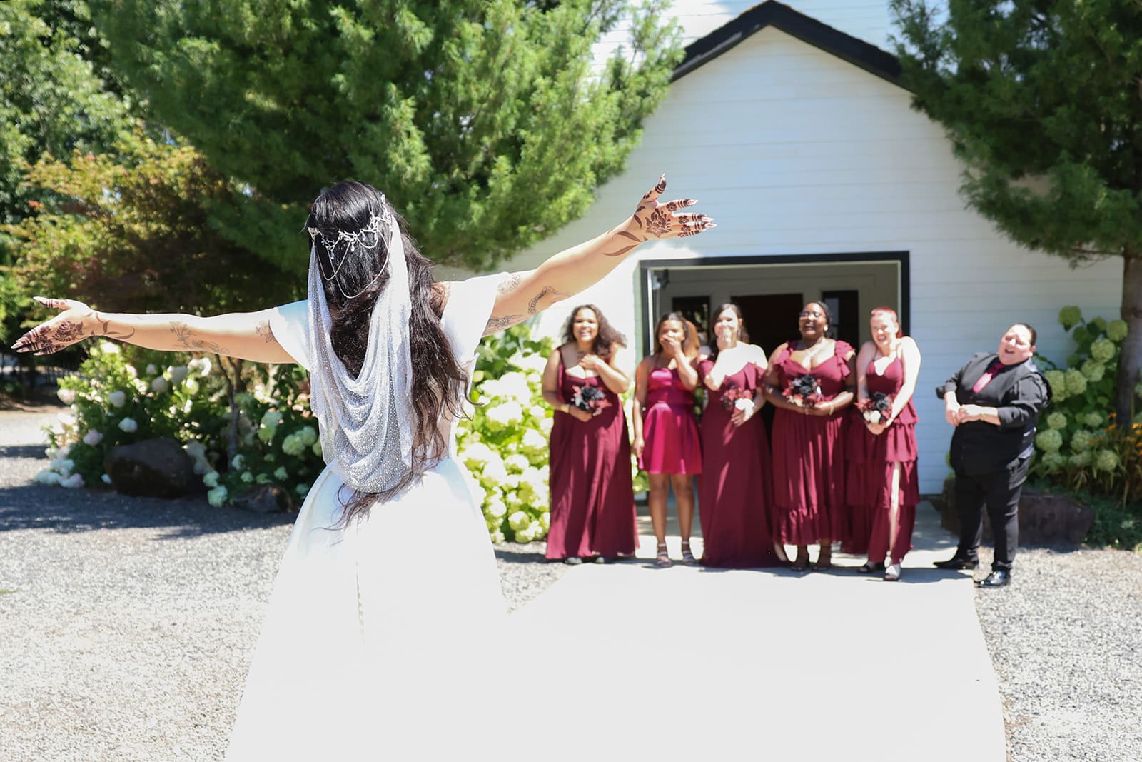 The bride, arms outstretched, walks toward her cheering bridal party during the first look outside the white chapel-style building at Cape Horn Estate.
