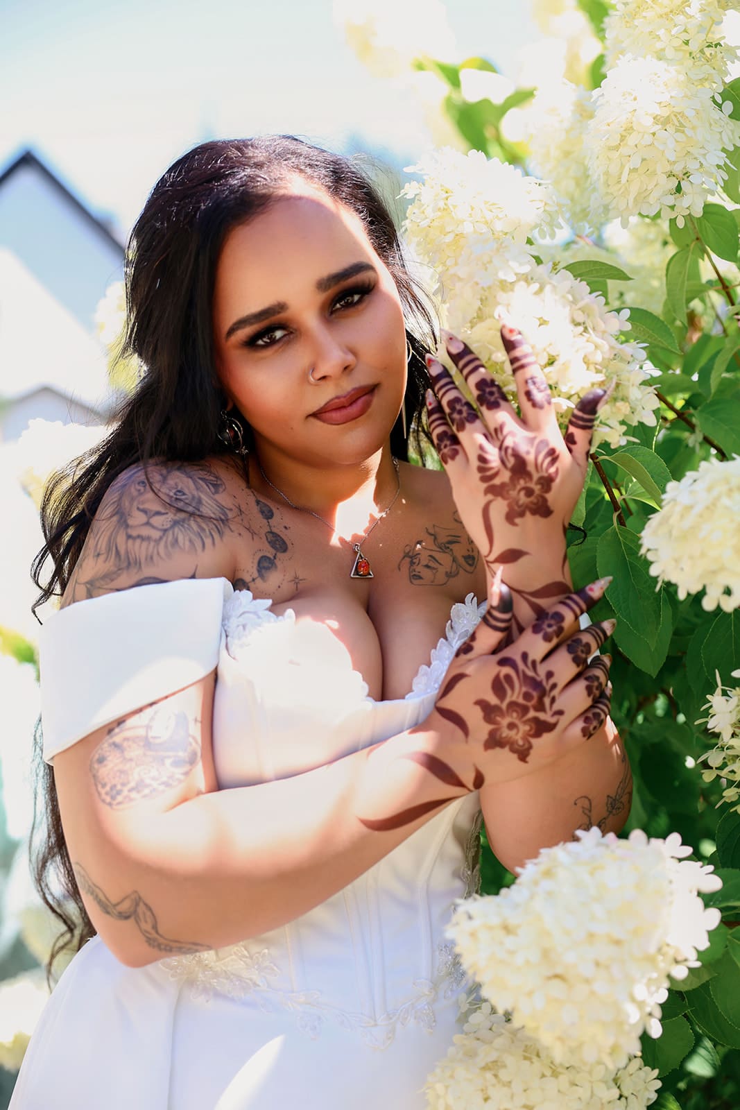 Bride poses among white hydrangeas, showing off intricate henna and tattoos in soft afternoon light.