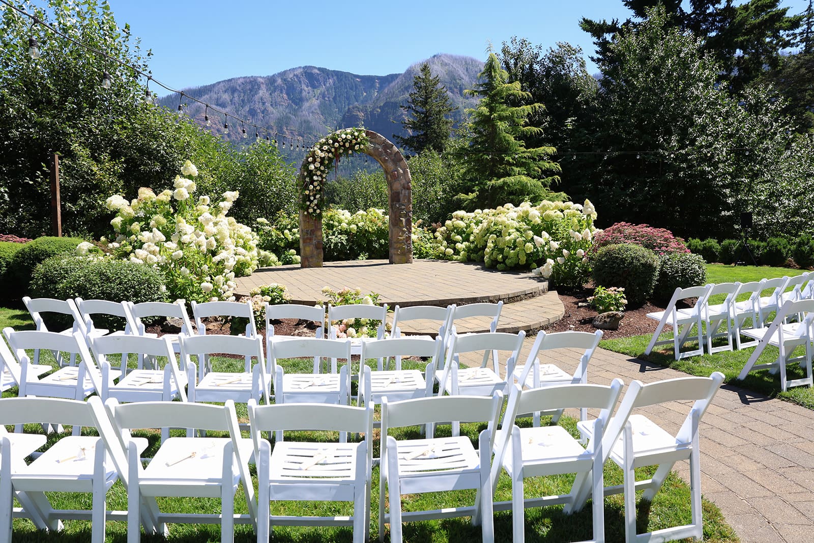 Empty white chairs face a round wooden ceremony platform with a floral stone arch, overlooking the Columbia River Gorge mountains at Cape Horn Estate in Oregon.