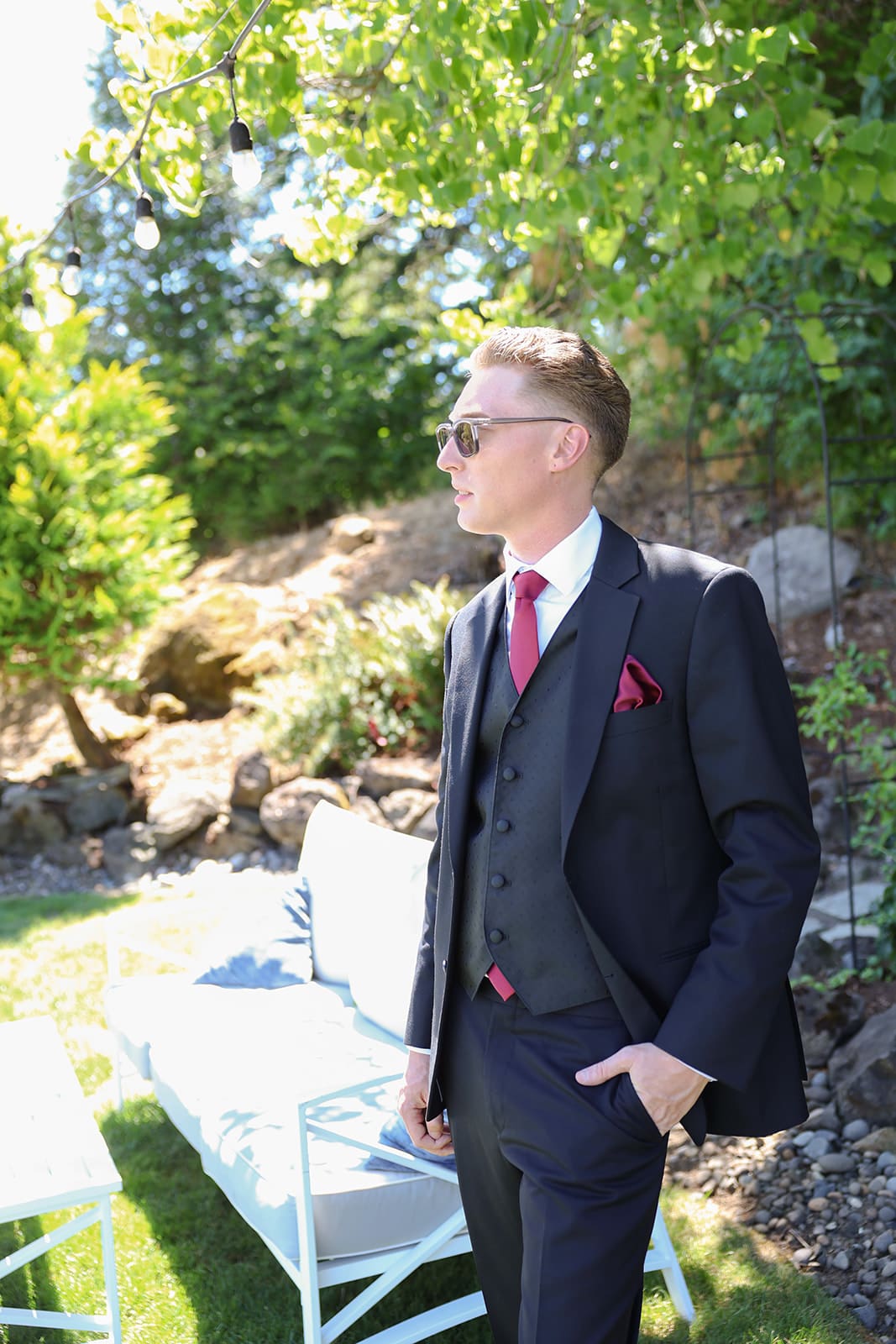 Groom stands in natural light under a tree canopy, wearing a black suit with burgundy details at an Oregon wedding venue.