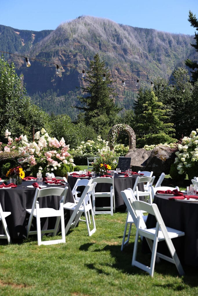 Round guest tables with black tablecloths and red napkins are set up on the lawn, framed by blooming florals and a dramatic mountain backdrop at an Oregon wedding venue.