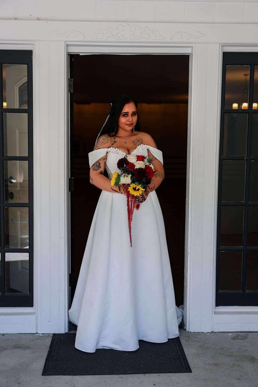 Portrait of the bride standing in the doorway of the reception building holding a bouquet of sunflowers and deep red blooms, framed by French doors at Cape Horn Estate.