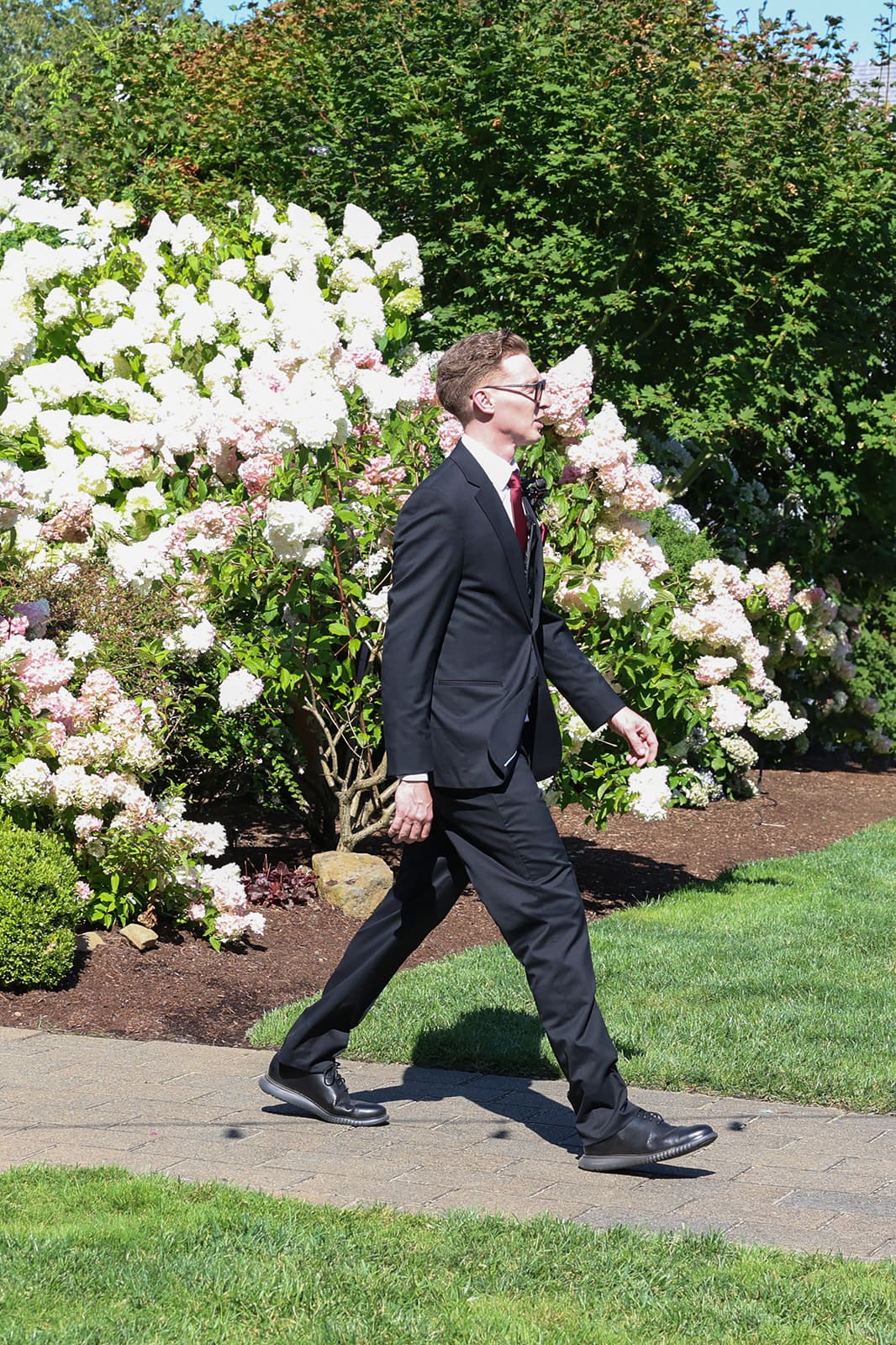 The groom walks along a landscaped path surrounded by blooming hydrangeas and manicured lawns on a sunny day at this peaceful Oregon wedding venue.