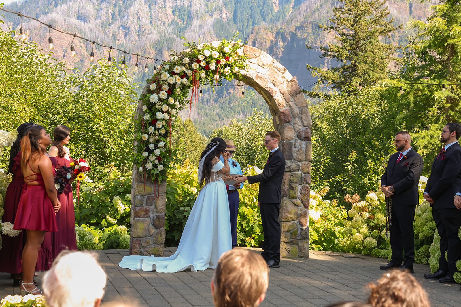 An outdoor wedding ceremony takes place under a flower-covered stone arch at Cape Horn Estate, with lush greenery and the Columbia Gorge cliffs rising behind the couple.