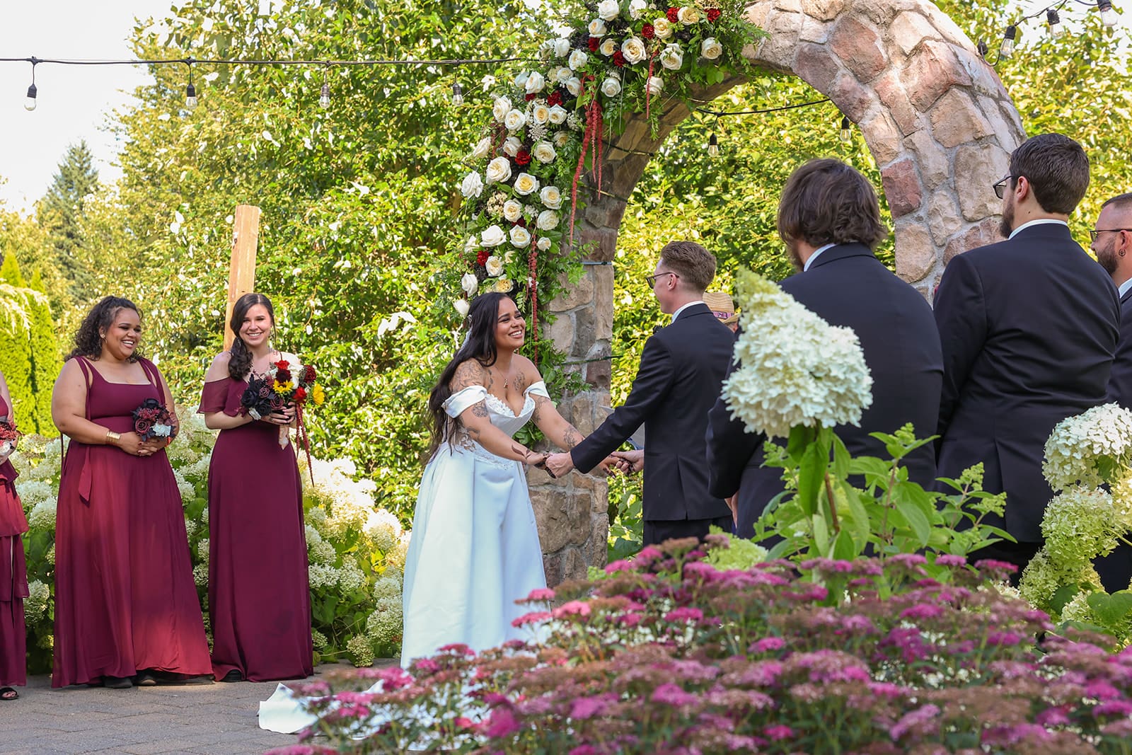 Bride and groom share a joyful moment during their ceremony under the floral-adorned stone arch, with bridesmaids in burgundy dresses smiling in the background at this scenic Oregon wedding venue.