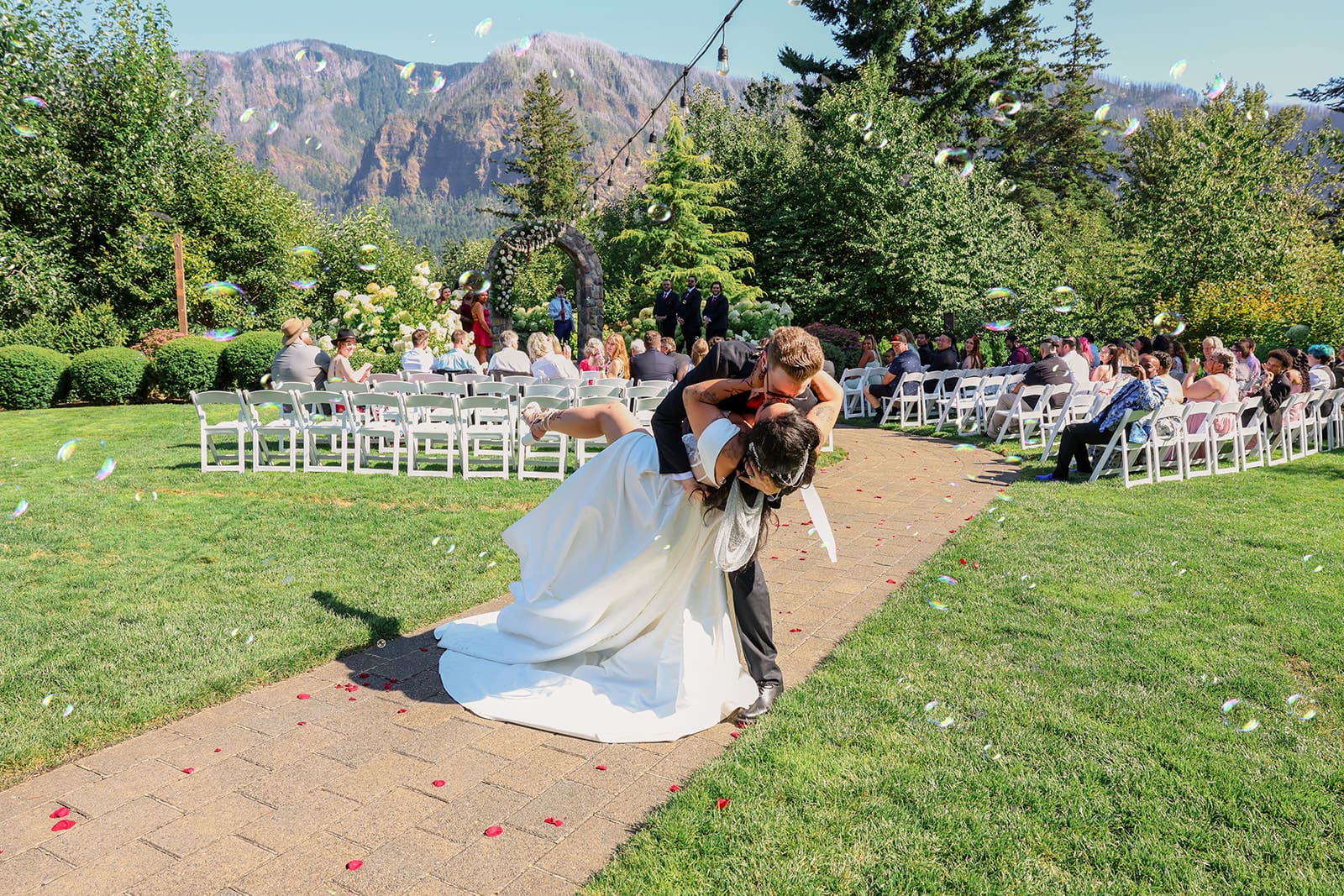 Groom dips bride for a kiss on the stone pathway after their ceremony at Cape Horn Estate, surrounded by white chairs and the dramatic mountain backdrop of this Oregon wedding venue.