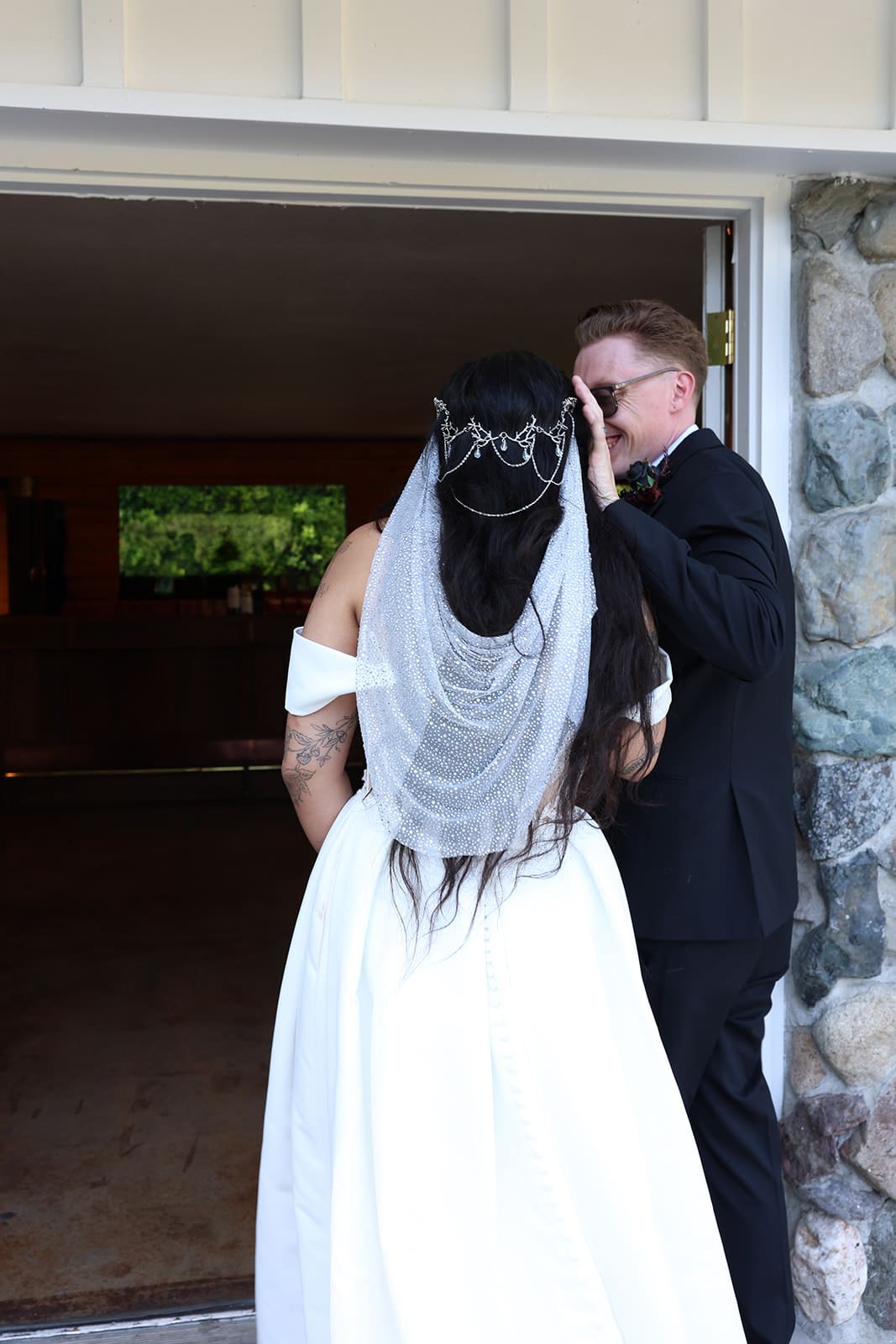 Bride and groom share a private moment before their ceremony, standing in a doorway with the bride’s detailed beaded veil and long dark hair cascading down her back. The groom, in a black suit, gently touches his face as they smile together.