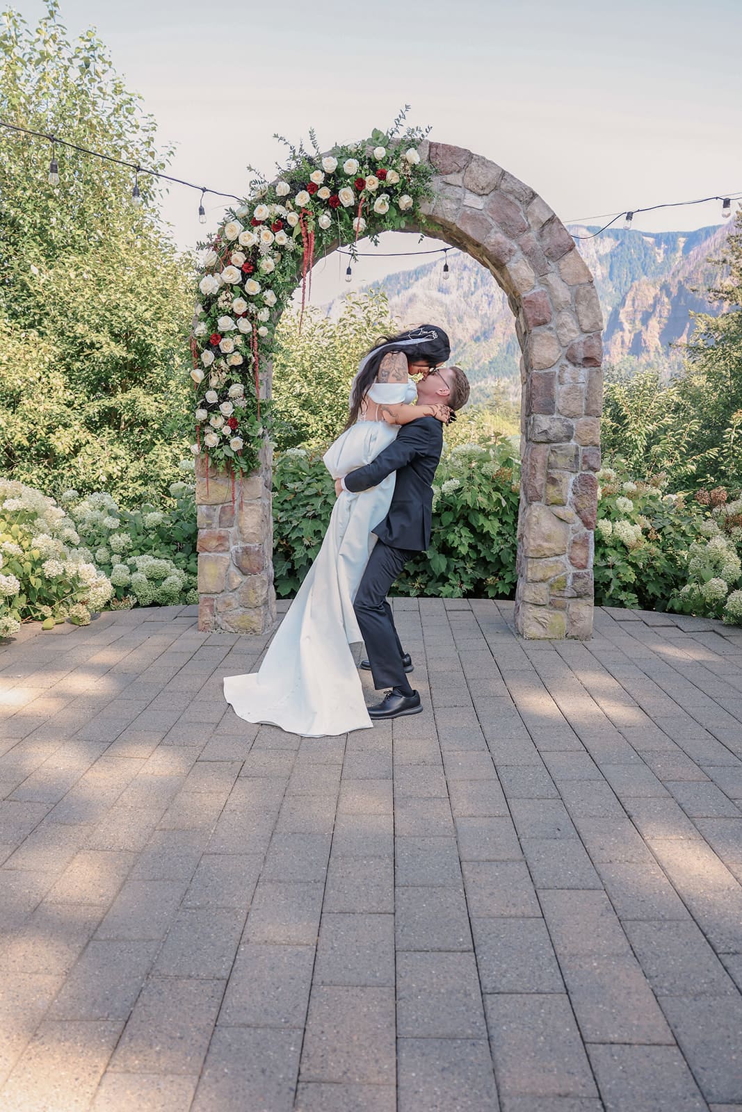 Full-length portrait of the couple sharing a kiss under a stone arch adorned with cream and rust roses at an Oregon wedding venue.