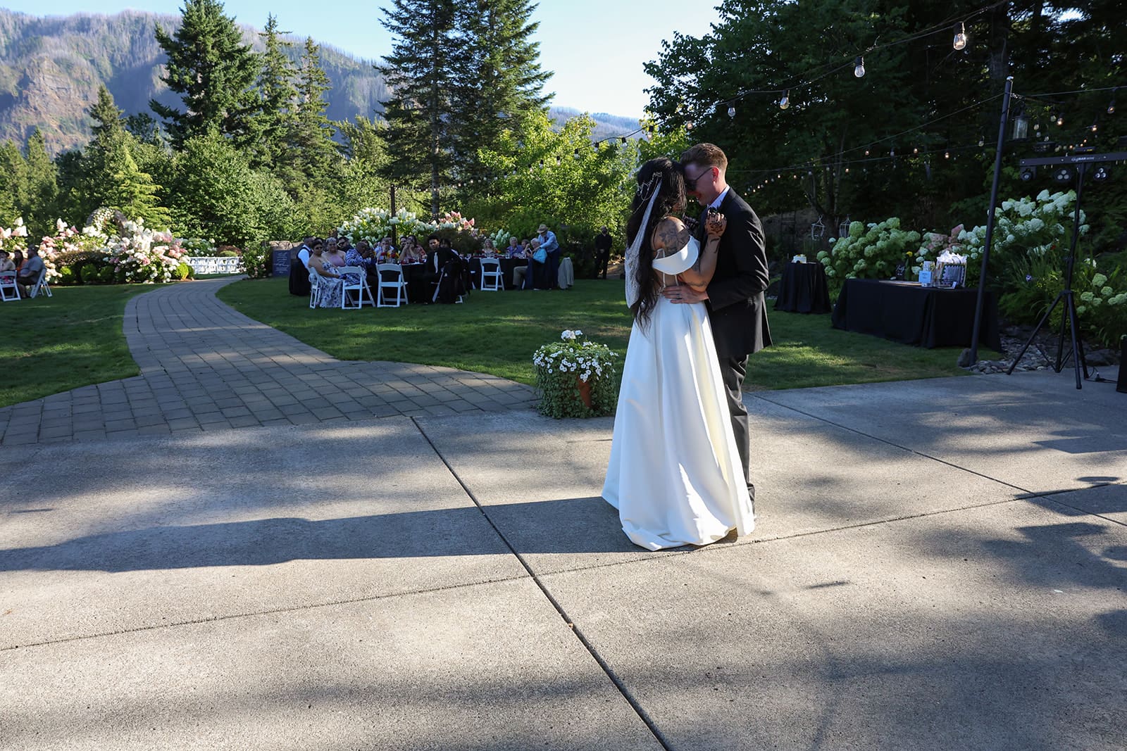 The newlyweds sway during their first dance on the outdoor patio, surrounded by lush greenery and mountain views at an Oregon wedding venue.