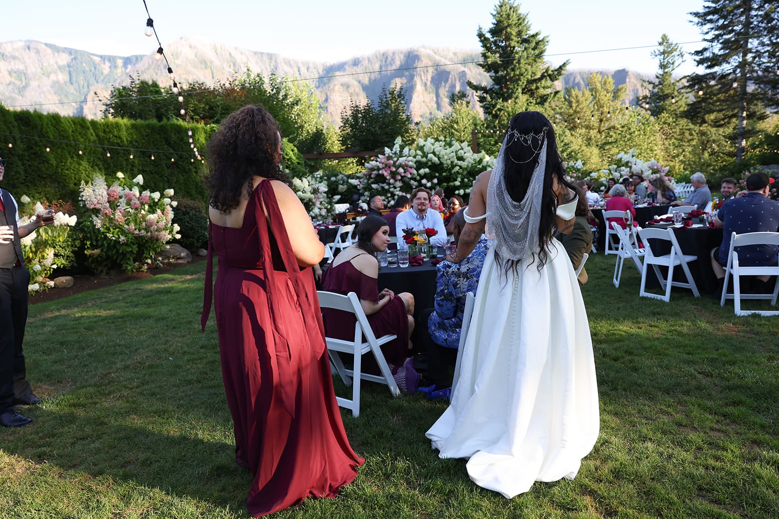 The bride and her bridesmaid walk through the lawn toward seated guests at a garden-style Oregon wedding venue with mountain views.