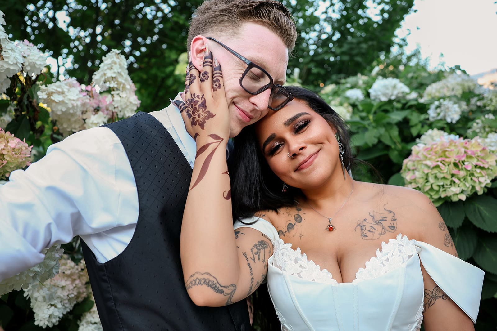 With her head nestled on his shoulder, the bride smiles contentedly as the groom wraps his arm around her, both framed by a backdrop of soft, blooming florals.