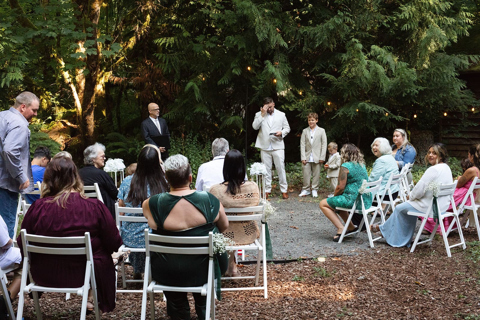 Guests gather outdoors among tall trees, seated in white chairs as the ceremony begins in an intimate backyard airbnb wedding.