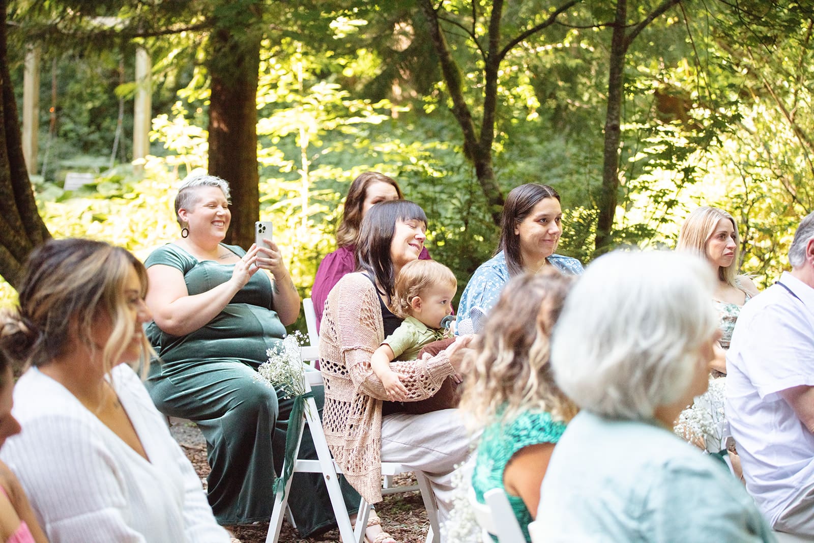 Guests laugh and smile from their seats during the ceremony, sunlight streaming through the forest canopy.