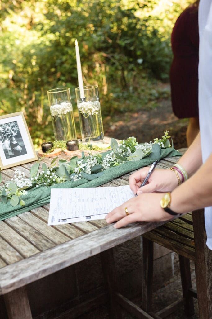 A close-up of a hand signing the marriage license on a wooden table covered with greenery and candles.