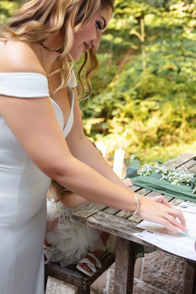 The bride signs the marriage license surrounded by greenery and soft golden light filtering through the forest.
