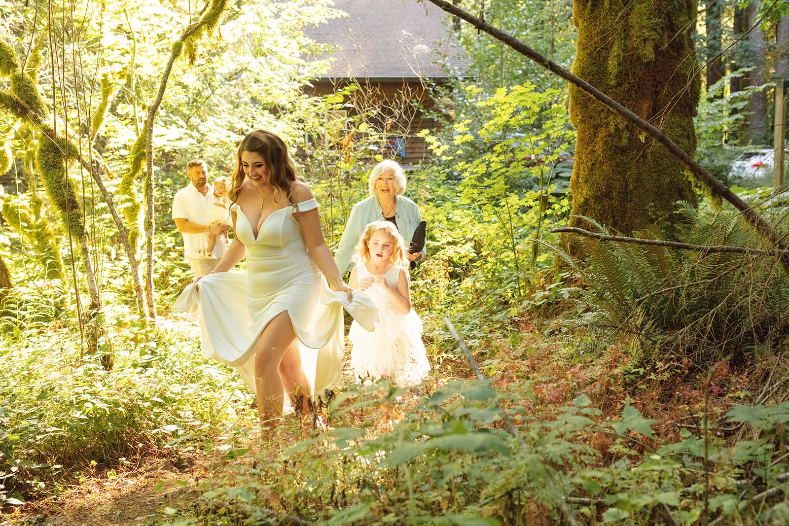 The bride walks through the woods with her daughter and grandmother, golden light filtering through the trees — a quintessential airbnb weddings moment.