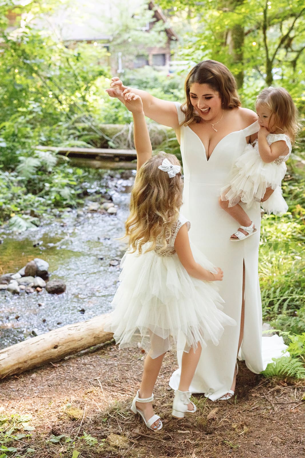 A bride in an off-shoulder gown twirls her two daughters in white dresses by a small creek, laughing together in the woods — the sweetest part of airbnb weddings.