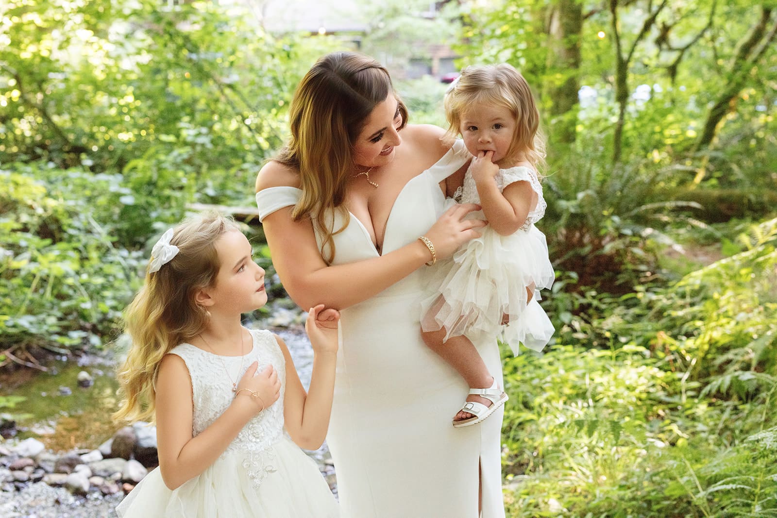 The bride shares a sweet moment with her two daughters in matching white dresses beside a forest creek — an airbnb weddings favorite.