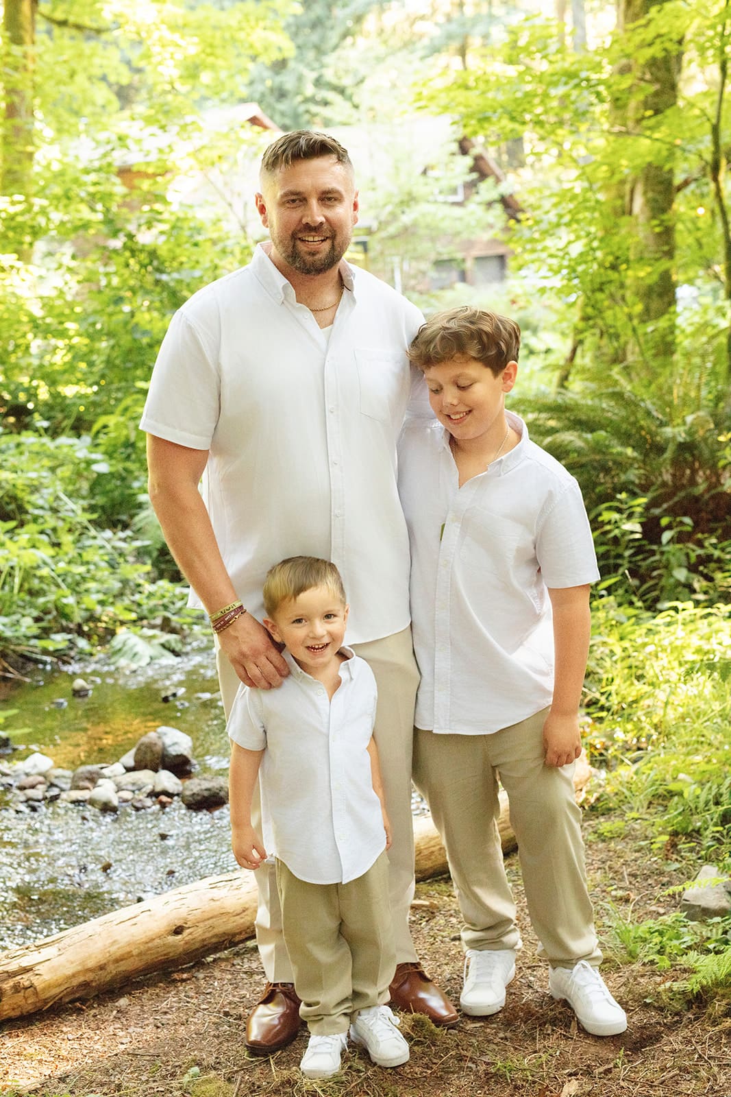 A groom poses with his sons by the creek, all wearing light neutral tones for a relaxed, summery feel.