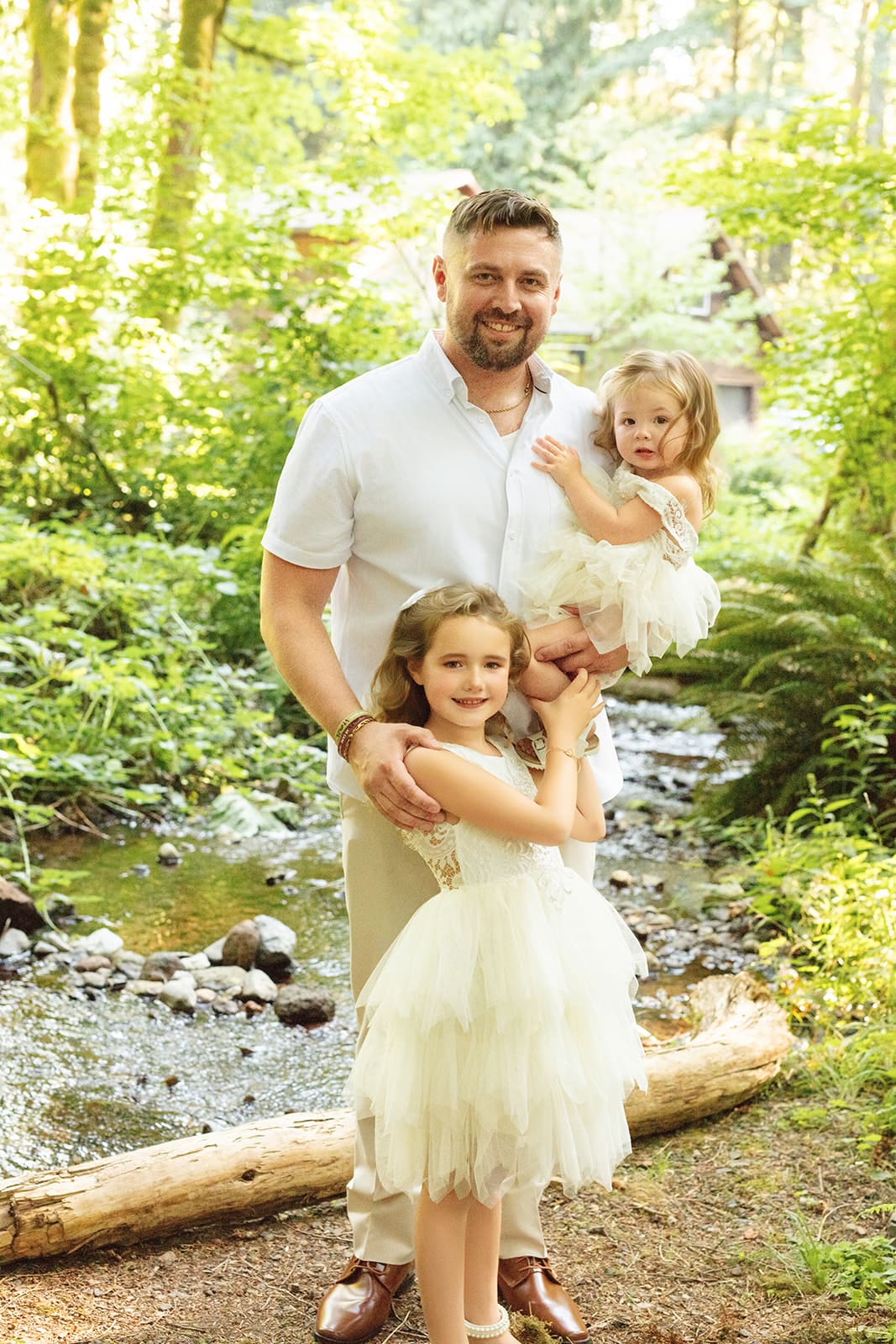 A groom smiles beside his two daughters near a forest creek, dressed in light tones that complement the natural backdrop.