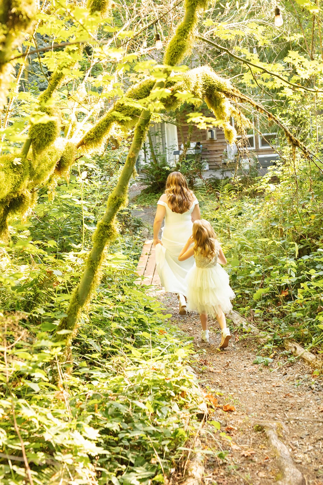 A bride and her daughter walk hand-in-hand down a mossy forest trail toward the cabin venue.