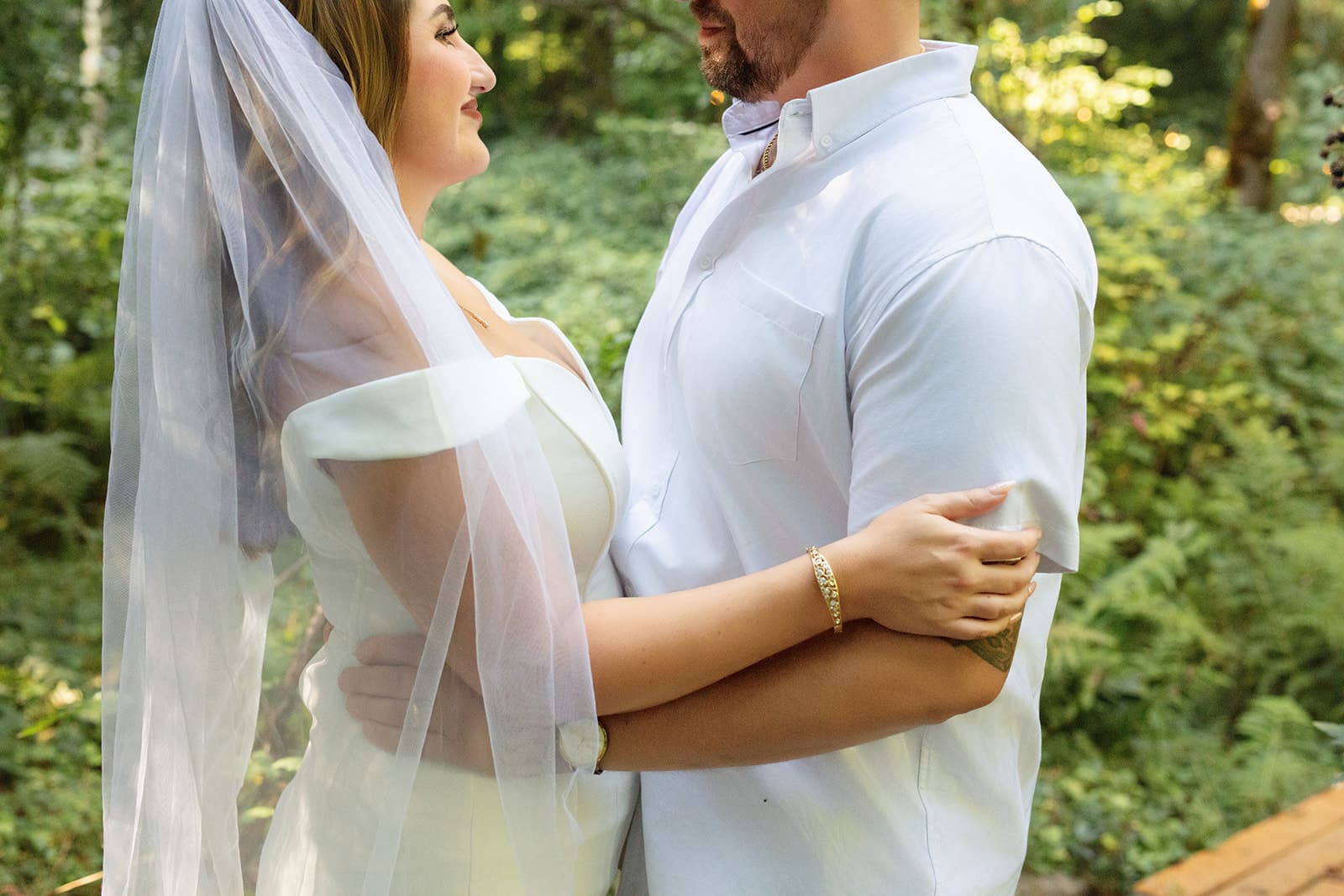 A close-up of the couple holding each other among lush forest greenery, sunlight softly filtering through the trees.