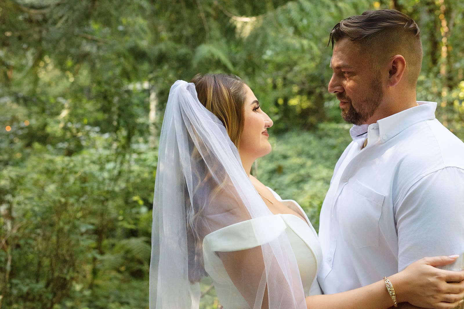 The couple stands close, smiling softly at each other as sunlight filters through the forest.