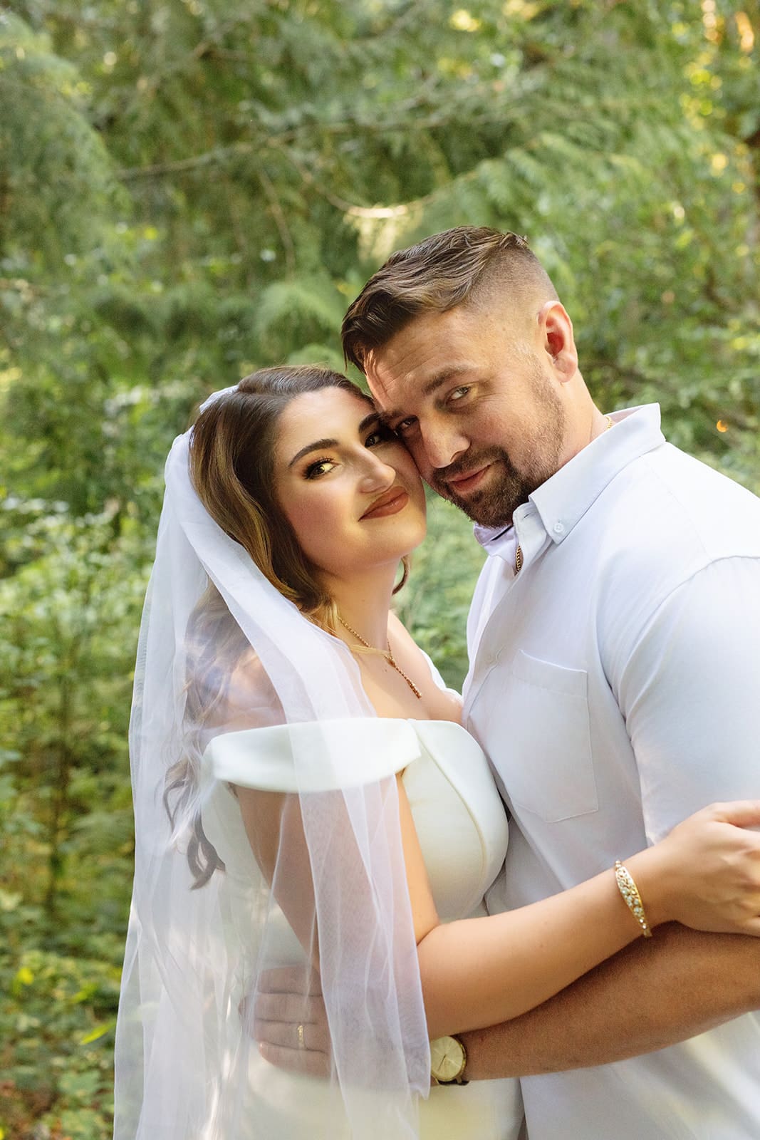 The couple embraces beneath the trees, her veil catching the light while he smiles down at her lovingly.