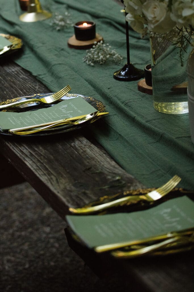 A rustic wooden table set with green linens, gold flatware, and black candleholders, styled for an intimate airbnb wedding dinner.
