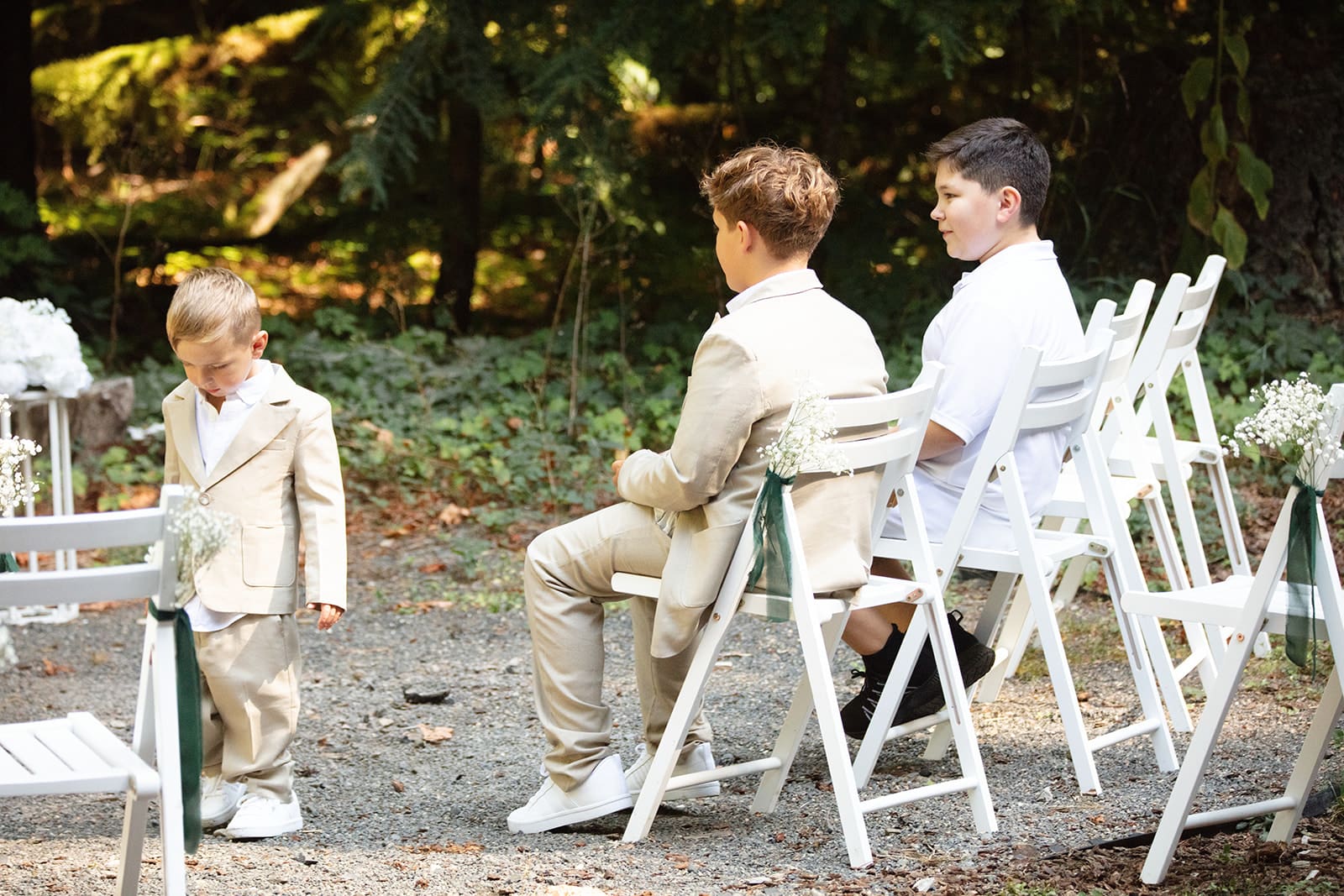 Three young boys in light suits and white shirts sit in folding chairs at an outdoor ceremony, waiting for the wedding to begin.