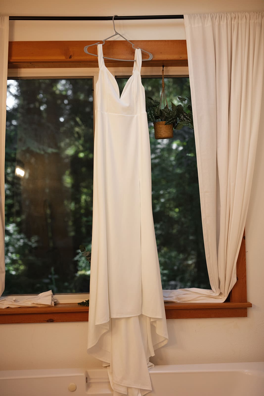 A minimalist white wedding dress hangs in front of a cabin window surrounded by forest light — a perfect setting for airbnb weddings.