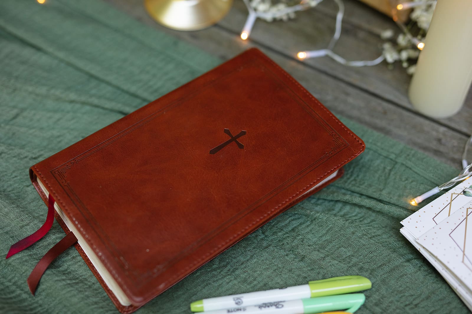 A brown leather Bible with a cross on the cover sits on a green linen cloth beside candles and string lights.