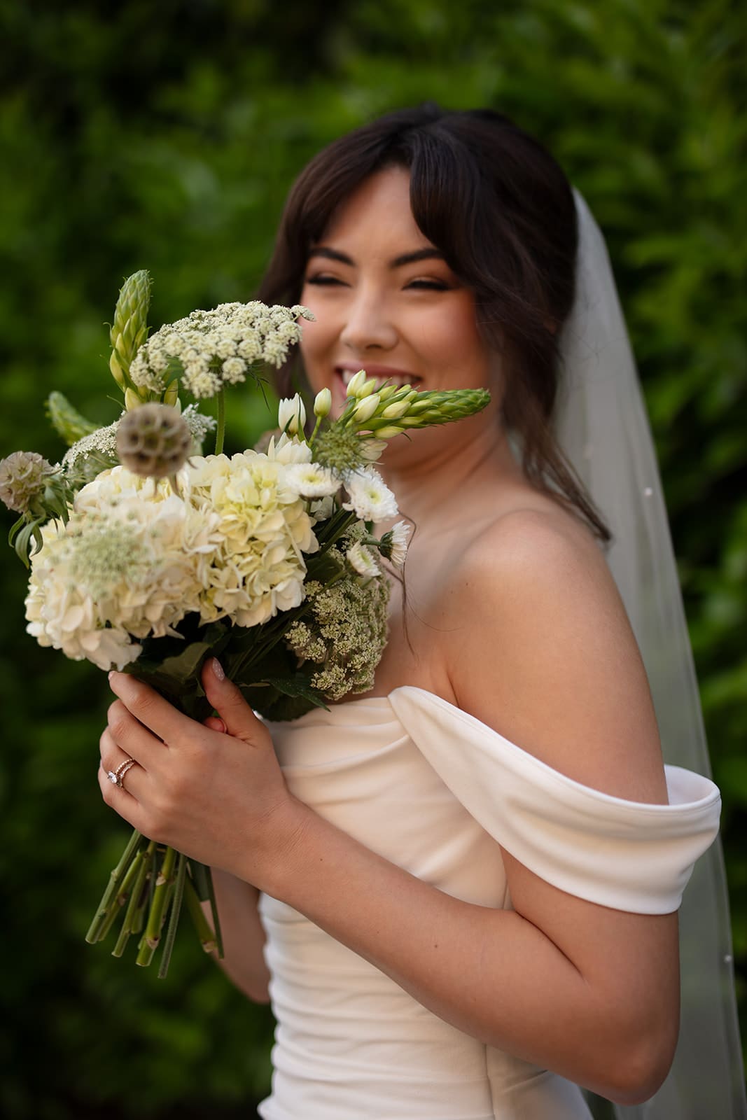 Radiant bride grinning behind her bouquet of white and green florals, full of pre-ceremony excitement.