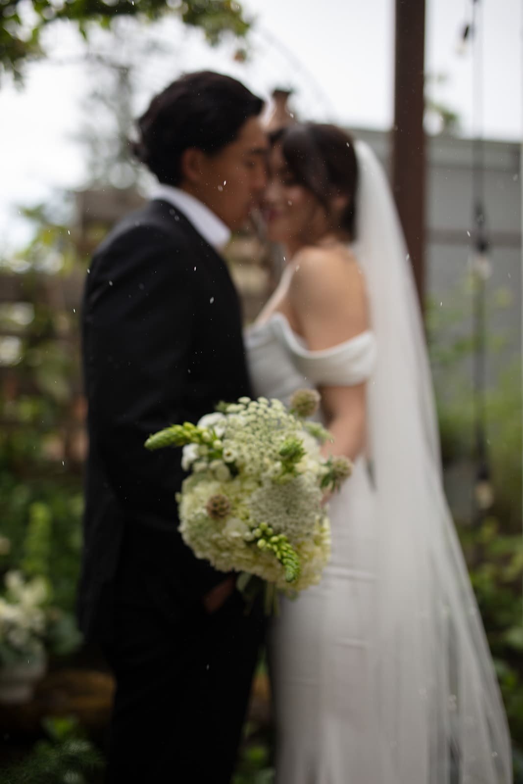 Intimate portrait with a blurred couple in the background and a textured bouquet in the foreground, highlighting the romantic tone of their celebration.