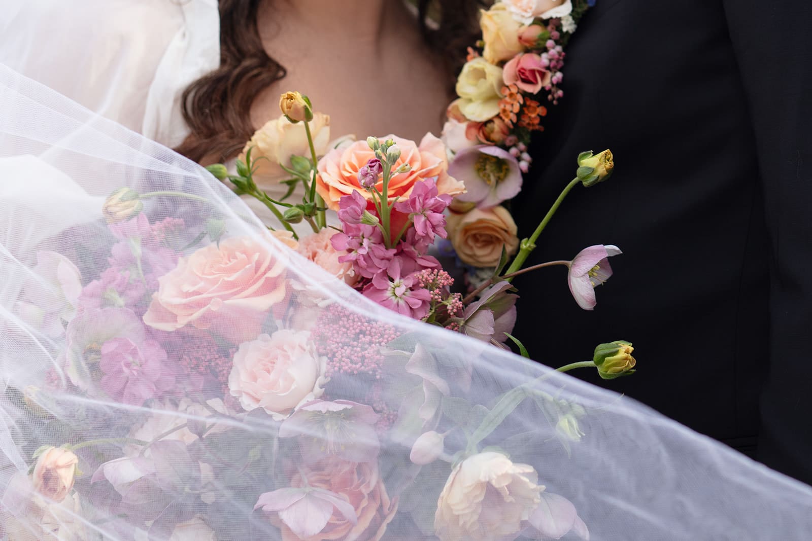 A layered floral arrangement featuring soft garden roses and wild textures, peeking through the bride’s veil.