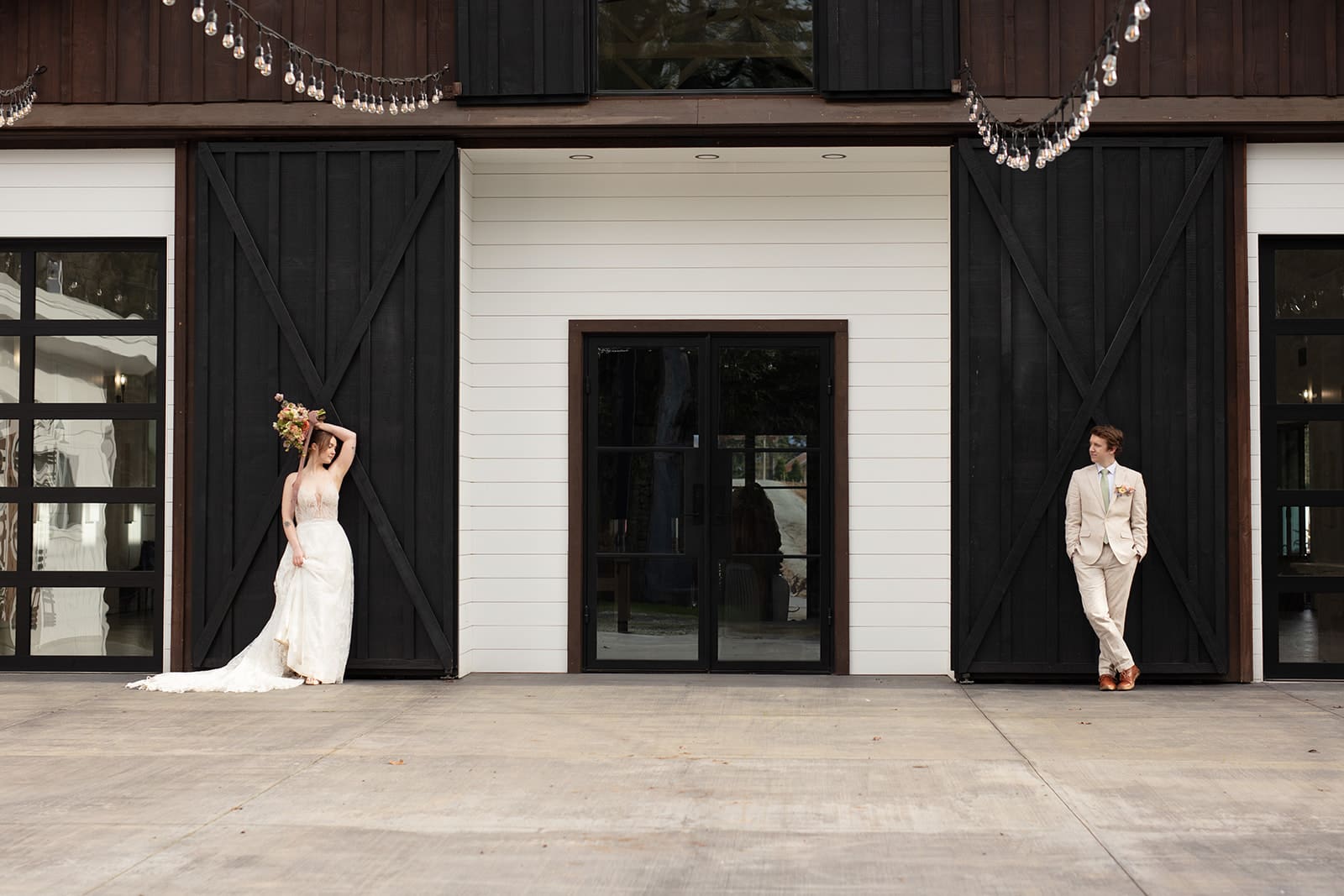 A stylish couple posing on opposite ends of a modern barn venue, capturing a fun wedding day moment.