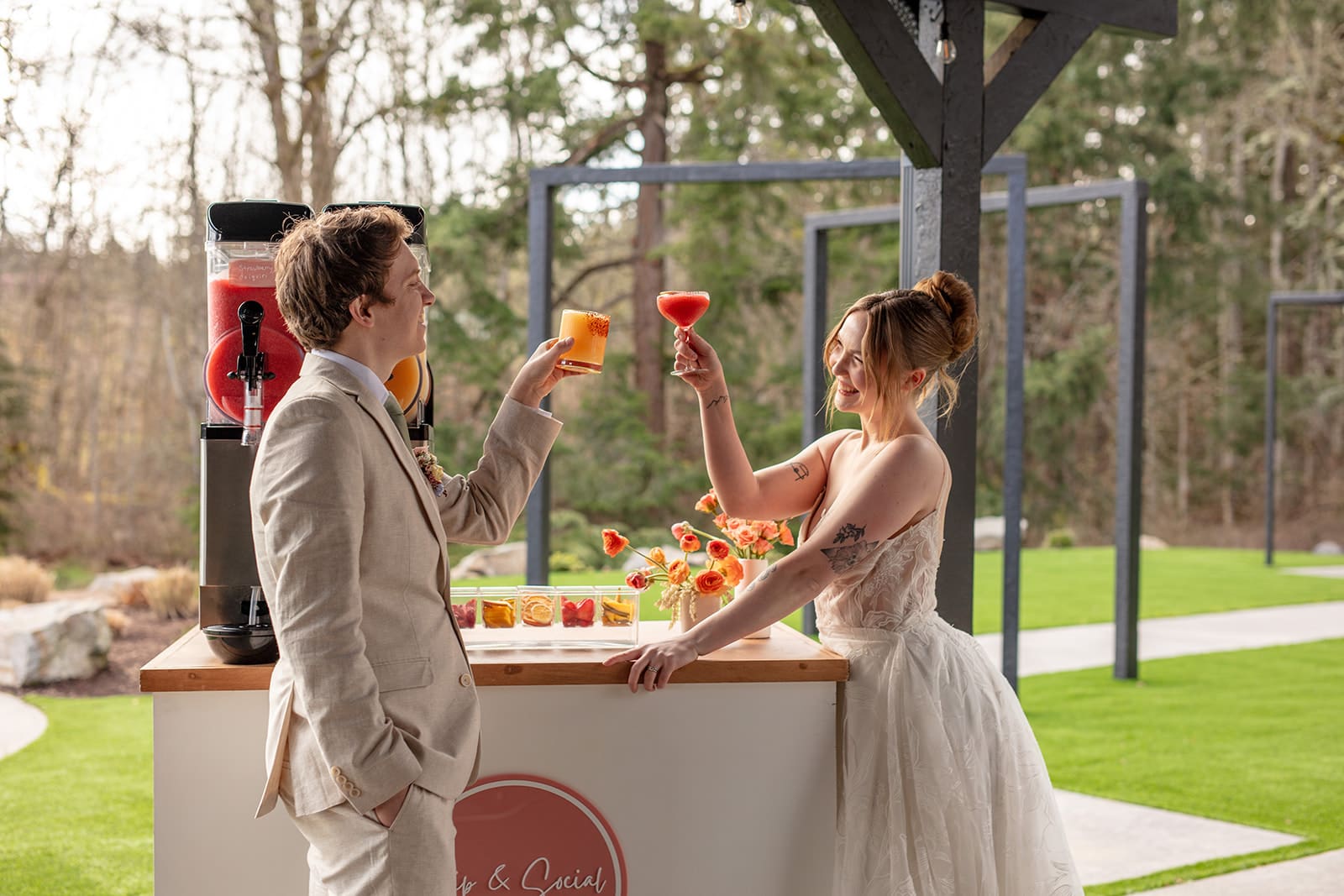 Bride and groom raising cocktails and smiling at a custom outdoor drink bar during their wedding celebration.